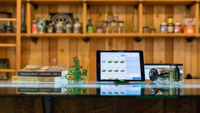 A tablet displaying a digital search interface for cannabis products, surrounded by glass items and books on a counter.